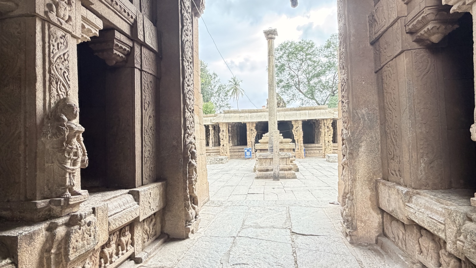 The view through a carved temple doorway into an open flagstone courtyard, a tall stone flag-pillar at its centre, pillared halls and a tree beyond.