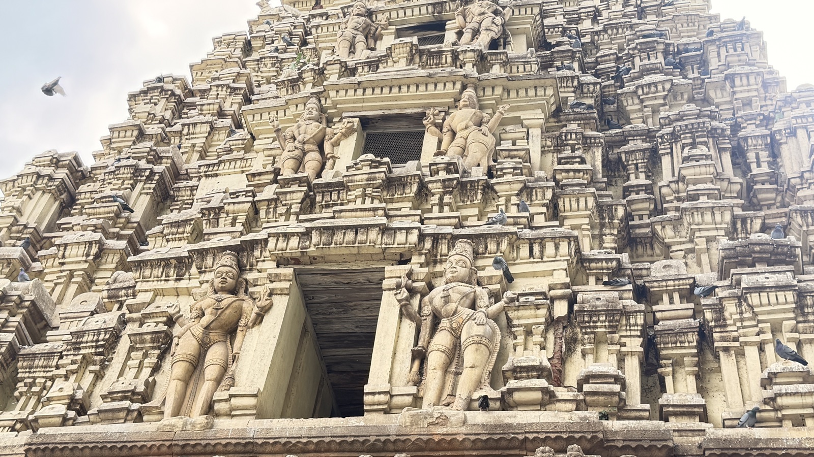 The lower tiers of a temple tower seen from below, two large sculpted guardian figures flanking a dark doorway, a bird caught mid-flight against the sky.